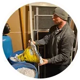 Man in a gray jacket and beanie pours salt from a bag into a blue container, near a water softener.
