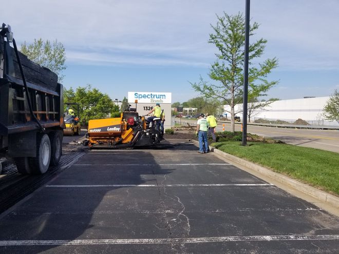 Road crew repairing asphalt on a parking lot, with a truck, orange paver, and workers on a sunny day