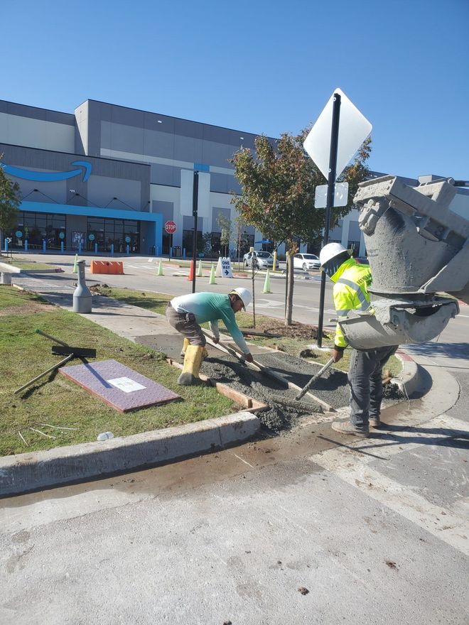 Workers repairing a sidewalk curb beside a building, with tools and a traffic sign nearby