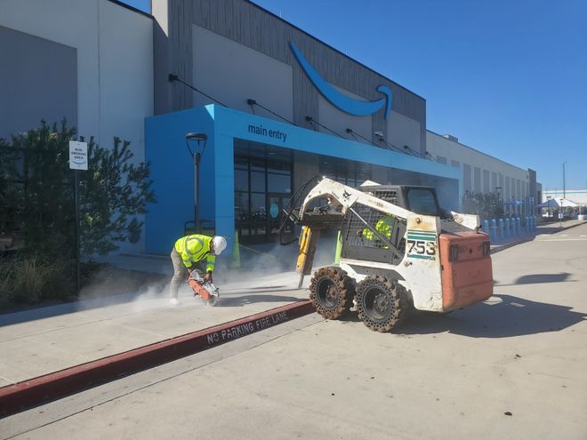 Workers operating a Bobcat cutting concrete outside a blue industrial building with dust spraying