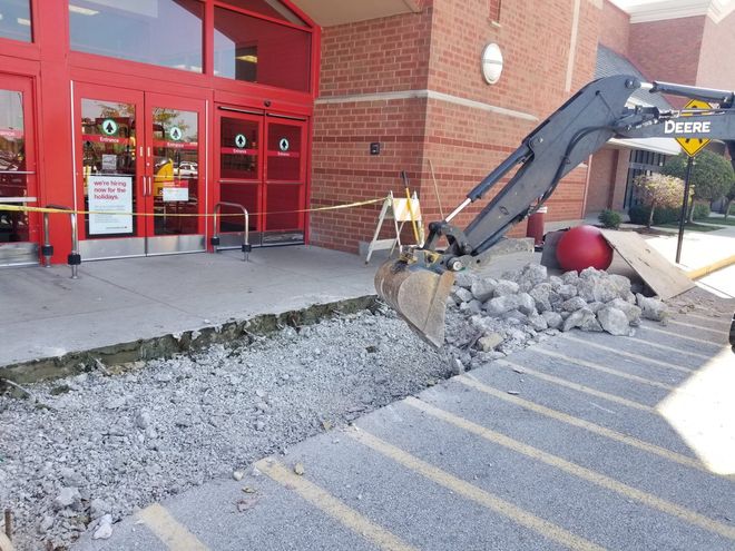 Excavator working beside a store entrance, removing gravel from a sidewalk and curb area