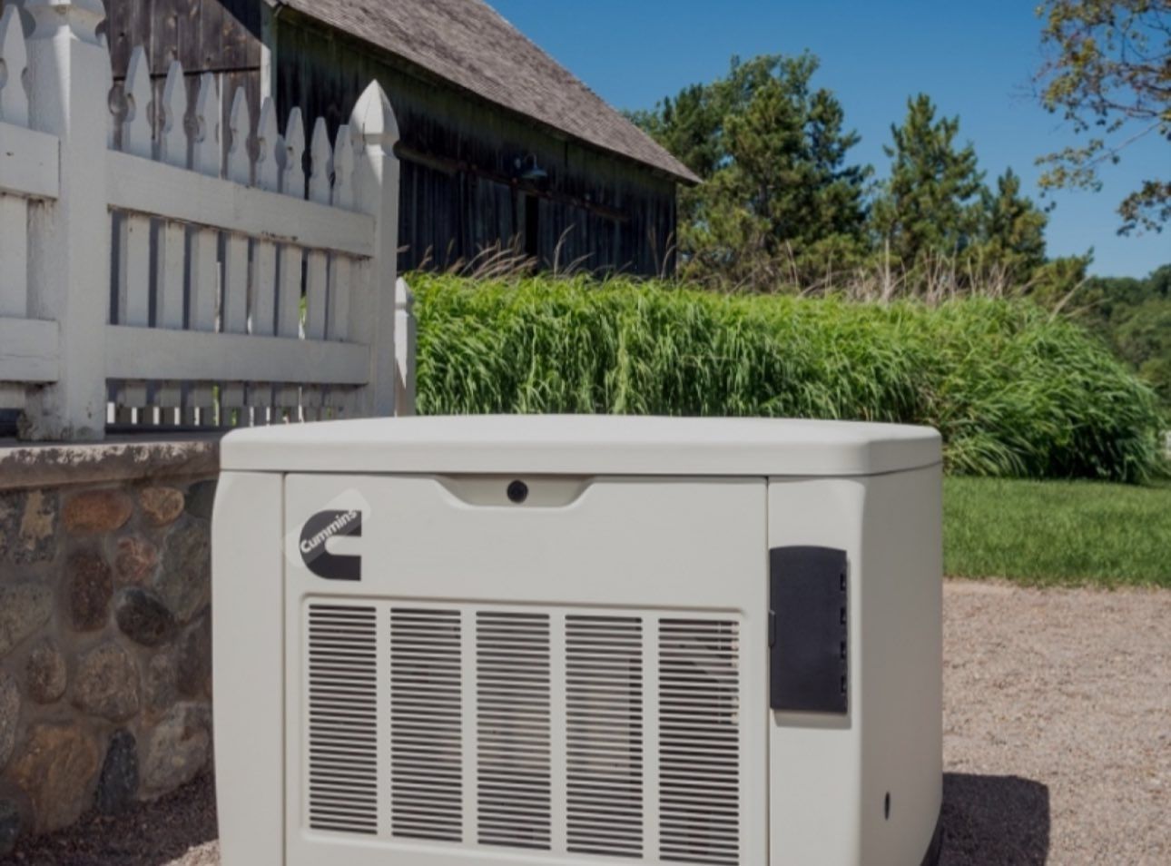 Cream-colored Cummins generator outside a white picket fence, barn, and greenery on a sunny day.