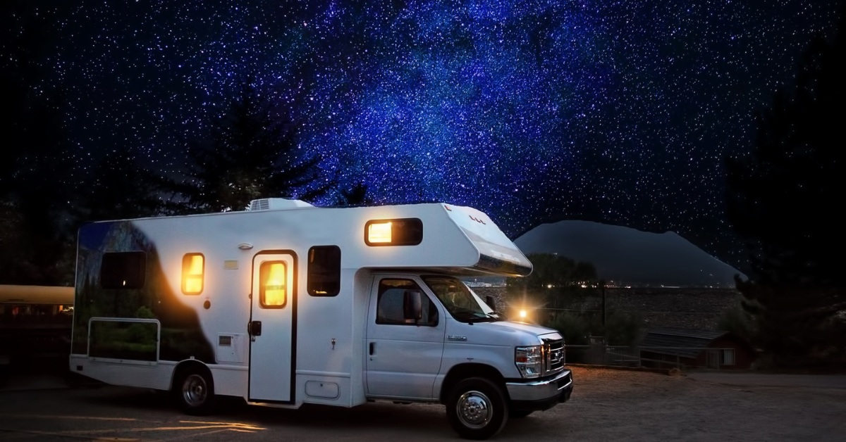 White RV parked under a starry night sky. Lit windows, dark landscape.