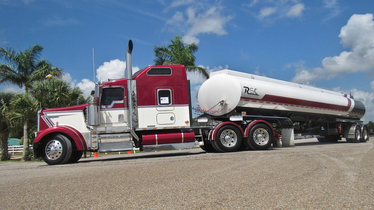 Red and white semi-truck tanker on gravel under a blue sky with palm trees.