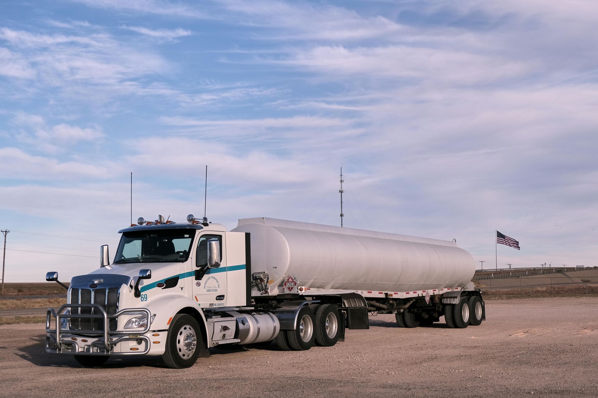 Semi-truck with a large silver tanker on a blue-sky day with a small American flag in the background.