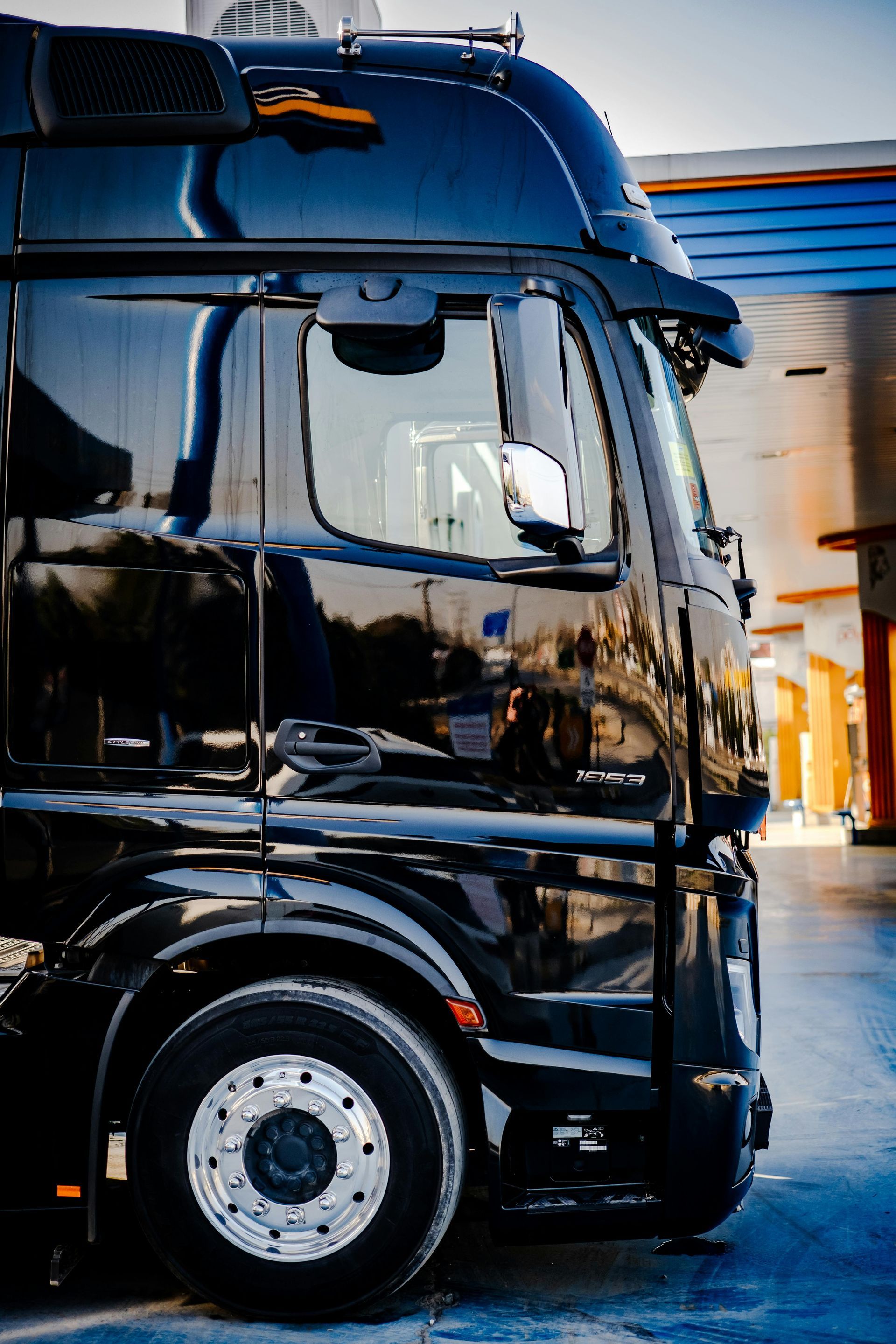 Black semi-truck parked at a gas station, side view. Shiny paint, chrome wheels, clear window reflection.