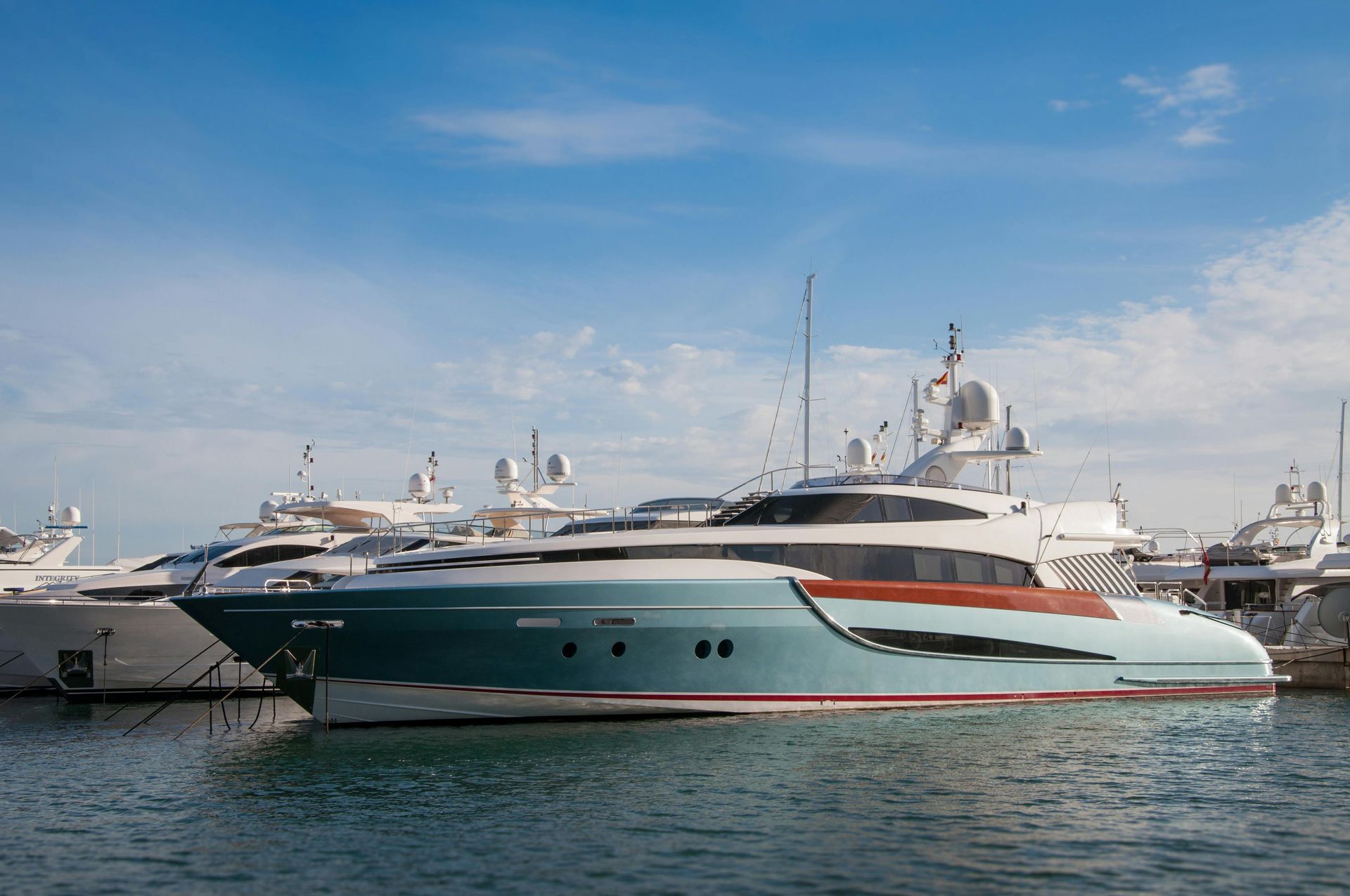 A teal and wood-trimmed yacht docked in a marina, with other yachts and a blue sky in the background.