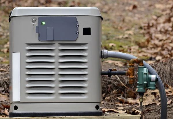 A gray home generator with a green indicator light, connected to a gas line, outdoors.