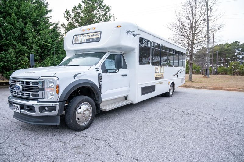 A white Ford shuttle bus parked on an asphalt lot with trees and a power pole in the background.