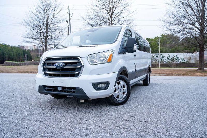 A white Ford Transit passenger van parked on an asphalt lot with trees in the background under a cloudy sky.