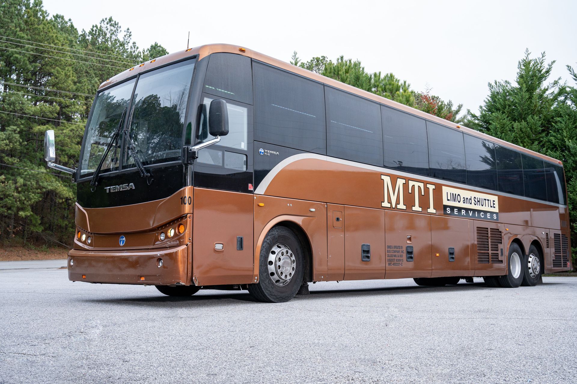 A bronze MTI motorcoach bus parked on a gravel lot in front of green trees.