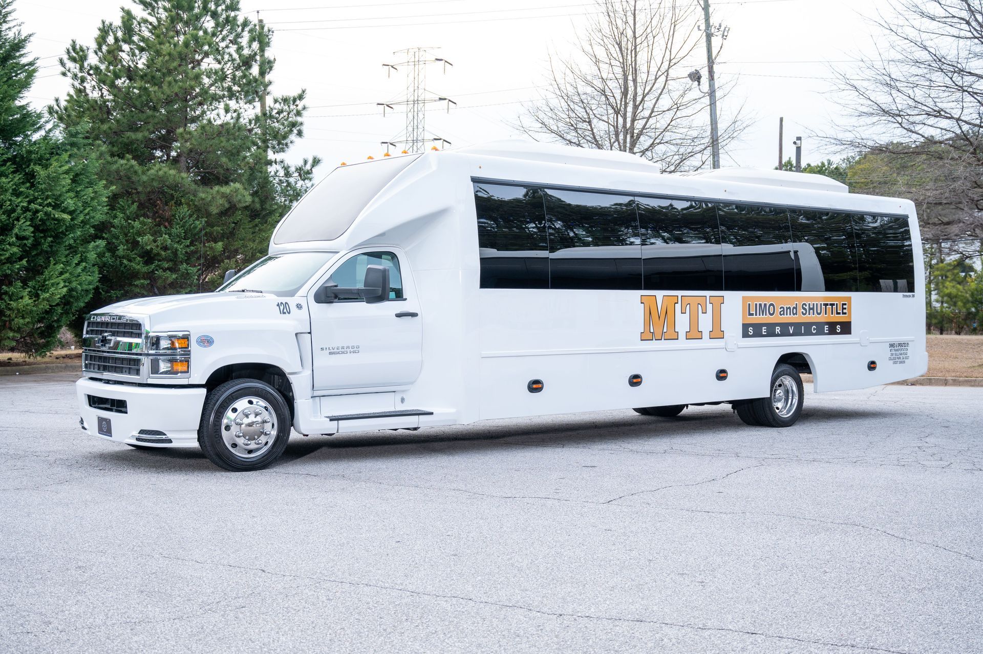 A white MTI party bus parked on a gravel surface outdoors with trees in the background.