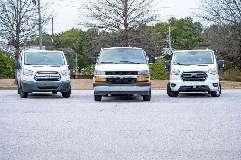 Three white cargo vans parked side-by-side on an asphalt lot with trees in the background.