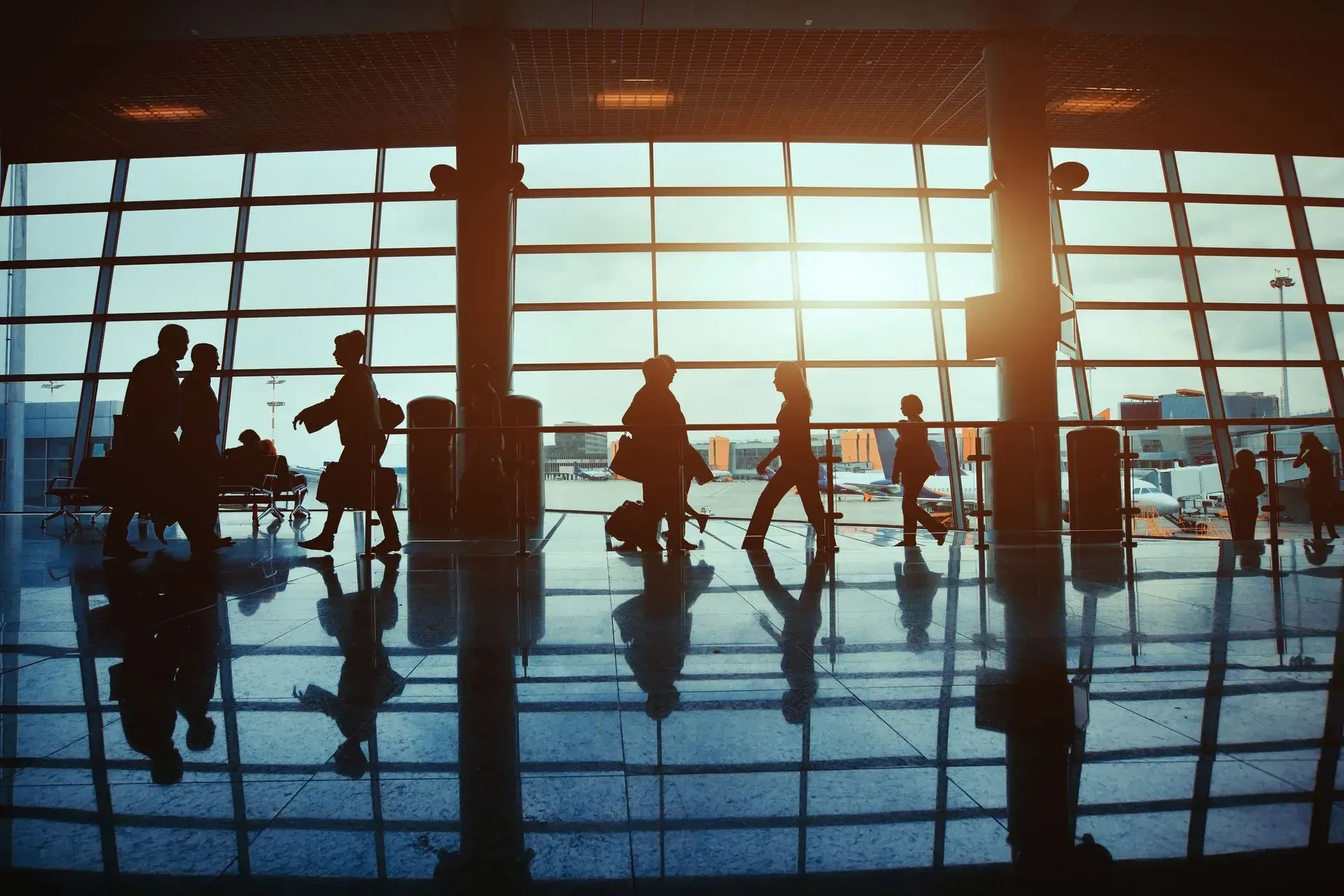 Silhouetted travelers walking through a sunlit airport terminal with reflective floors and large windows