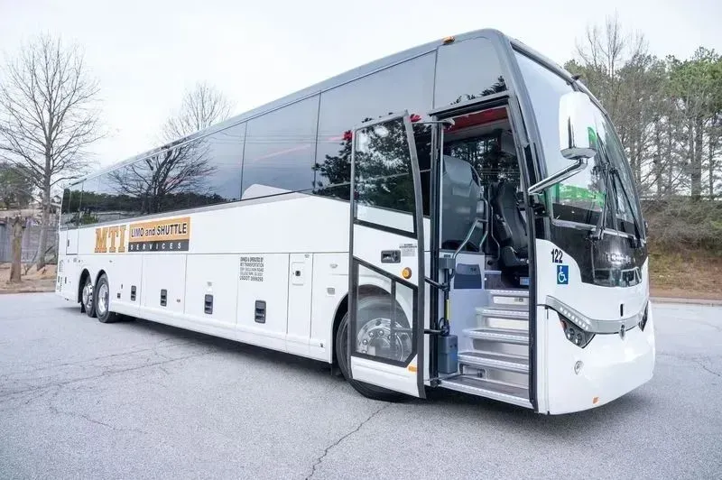 White coach bus parked outdoors with the front door open on a wet lot.