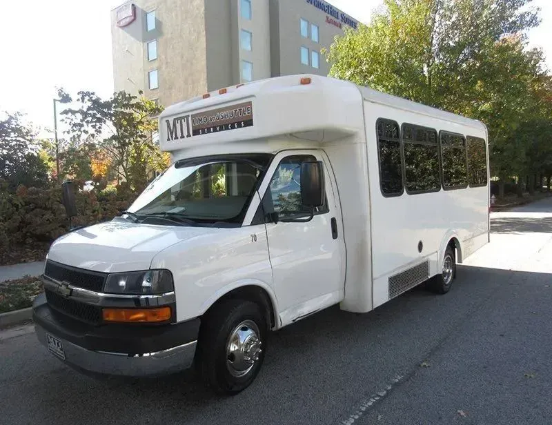 A white shuttle bus parked on a street beside apartment buildings and trees.