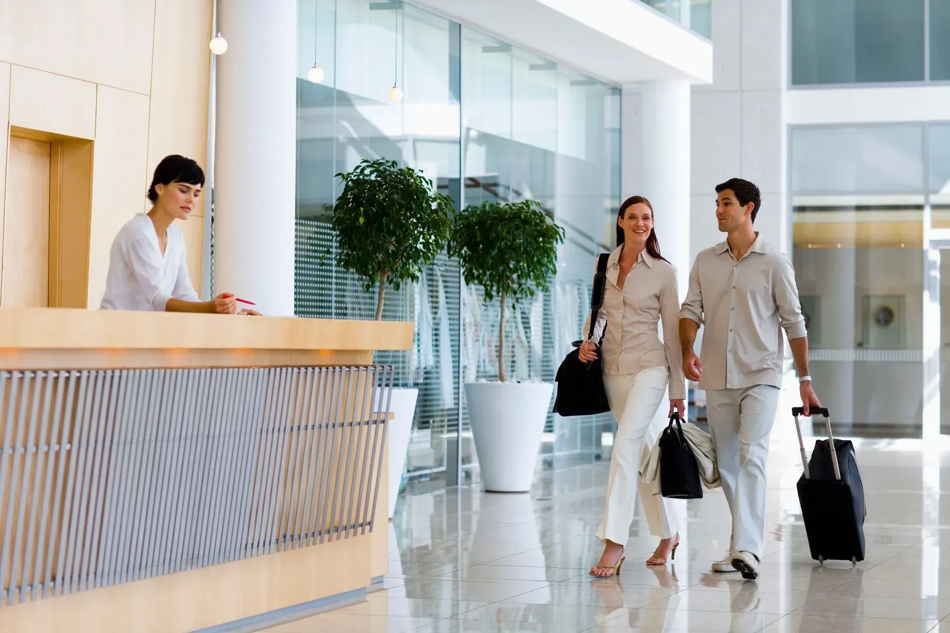 Two travelers with rolling suitcases walk through a bright hotel lobby past the front desk.