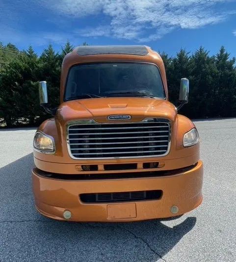A front view of an orange Freightliner semi-truck parked on an asphalt lot with a line of trees against a blue sky.
