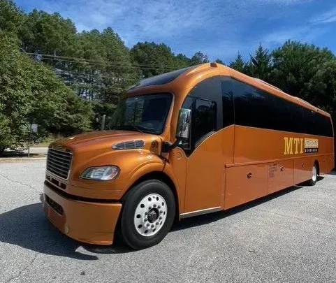 Orange and black MTA shuttle bus parked on a road beside trees under a blue sky