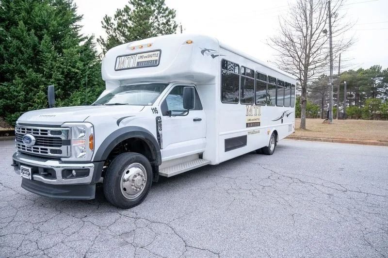 A white Ford shuttle bus parked on an asphalt lot with trees in the background.