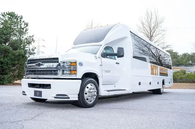 White luxury bus parked outdoors on a paved lot near trees and bushes