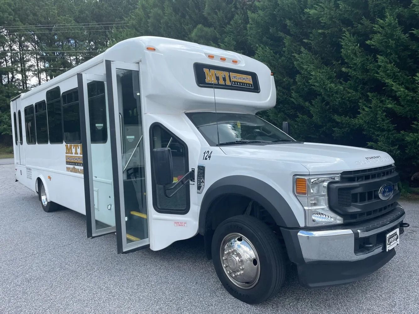 A white shuttle bus with a Ford chassis and an open passenger door parked on a gravel lot in front of trees.