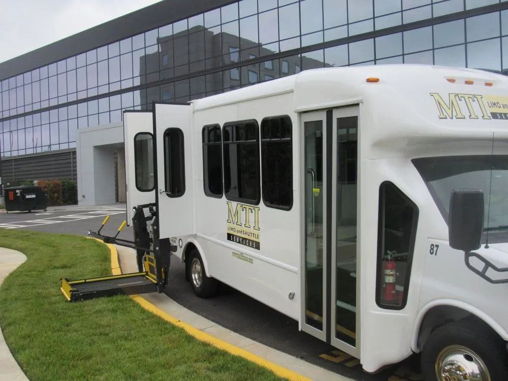 White wheelchair-accessible shuttle bus with its side lift deployed, parked on an asphalt driveway near a glass office building.