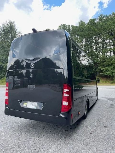 A rear view of a black shuttle bus parked on an asphalt surface with trees in the background under a cloudy blue sky.