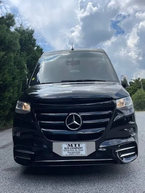 A front-facing black Mercedes-Benz Sprinter van parked on an asphalt surface under a cloudy sky.