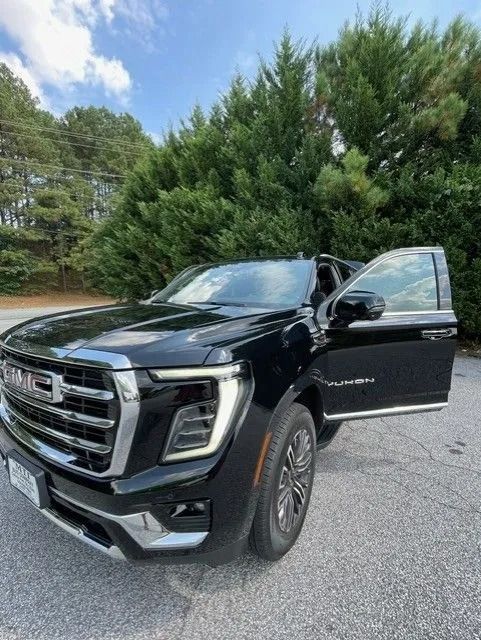 A black GMC Yukon SUV parked on asphalt with the front passenger-side door open, in front of pine trees.