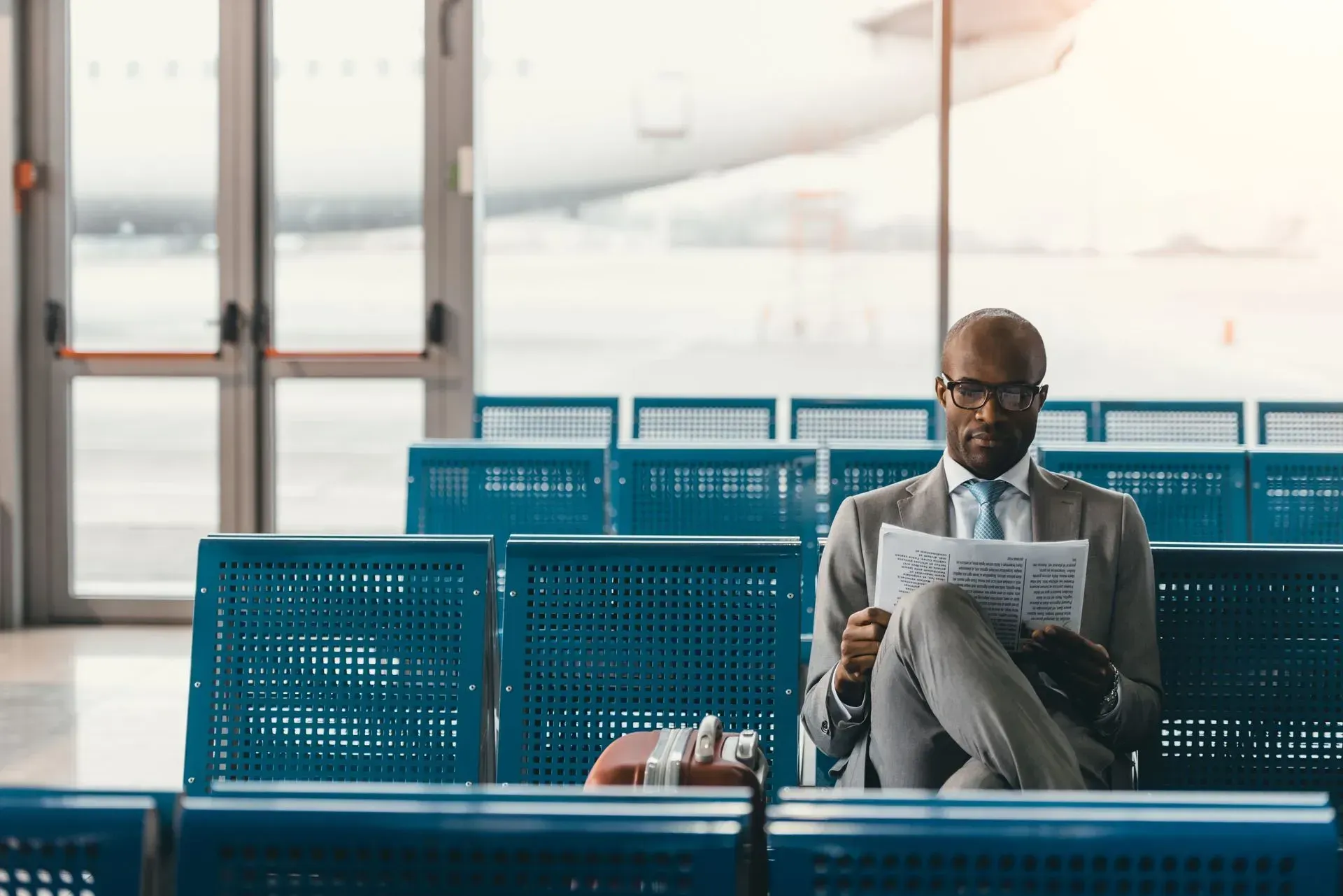 Man in glasses reading a newspaper in a bright airport waiting area with blue seats
