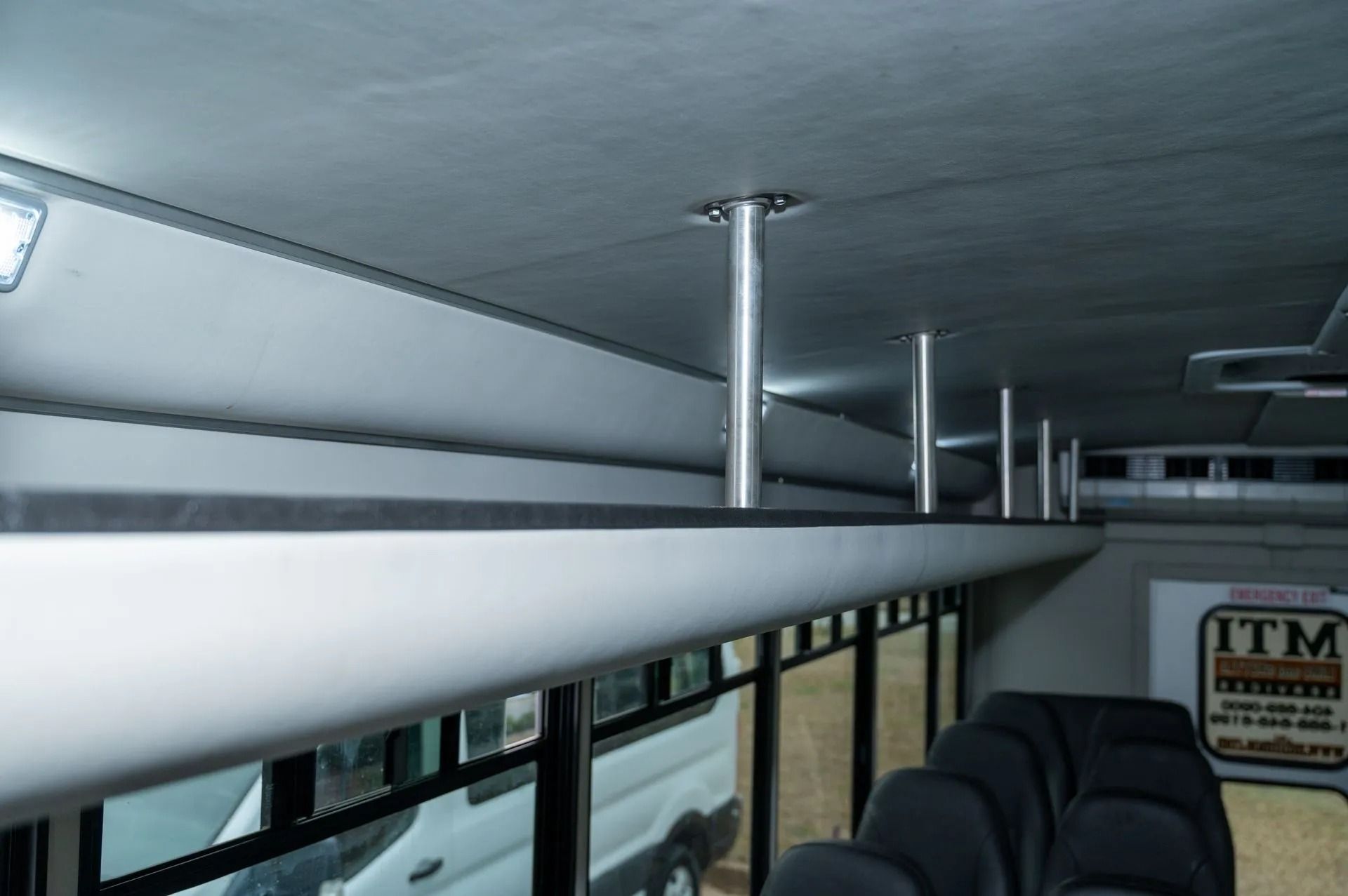 Interior view of a passenger bus showing gray overhead luggage racks supported by metal poles and gray seating.