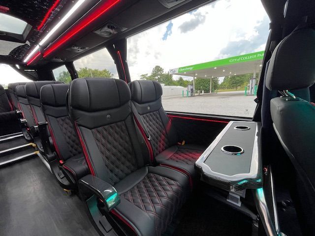 Interior of a luxury bus with black quilted leather seats, red accents, and a table with cup holders next to a window.