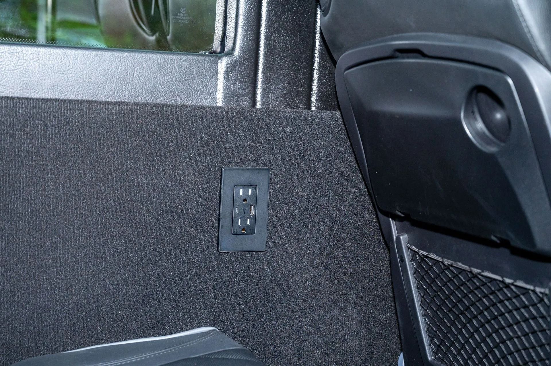 A standard electrical outlet mounted on a textured, dark gray upholstered panel inside a vehicle.