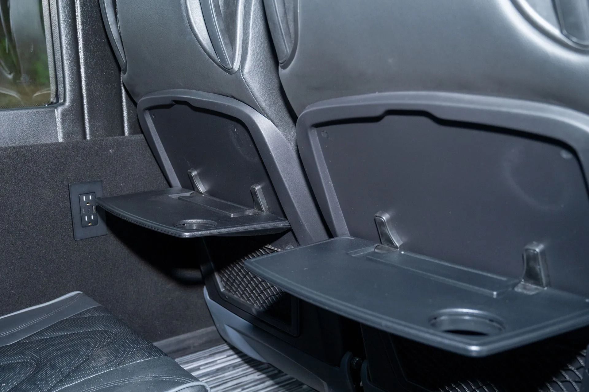 Interior view of bus seats featuring fold-down black tray tables, cup holders, and an accessible power outlet.