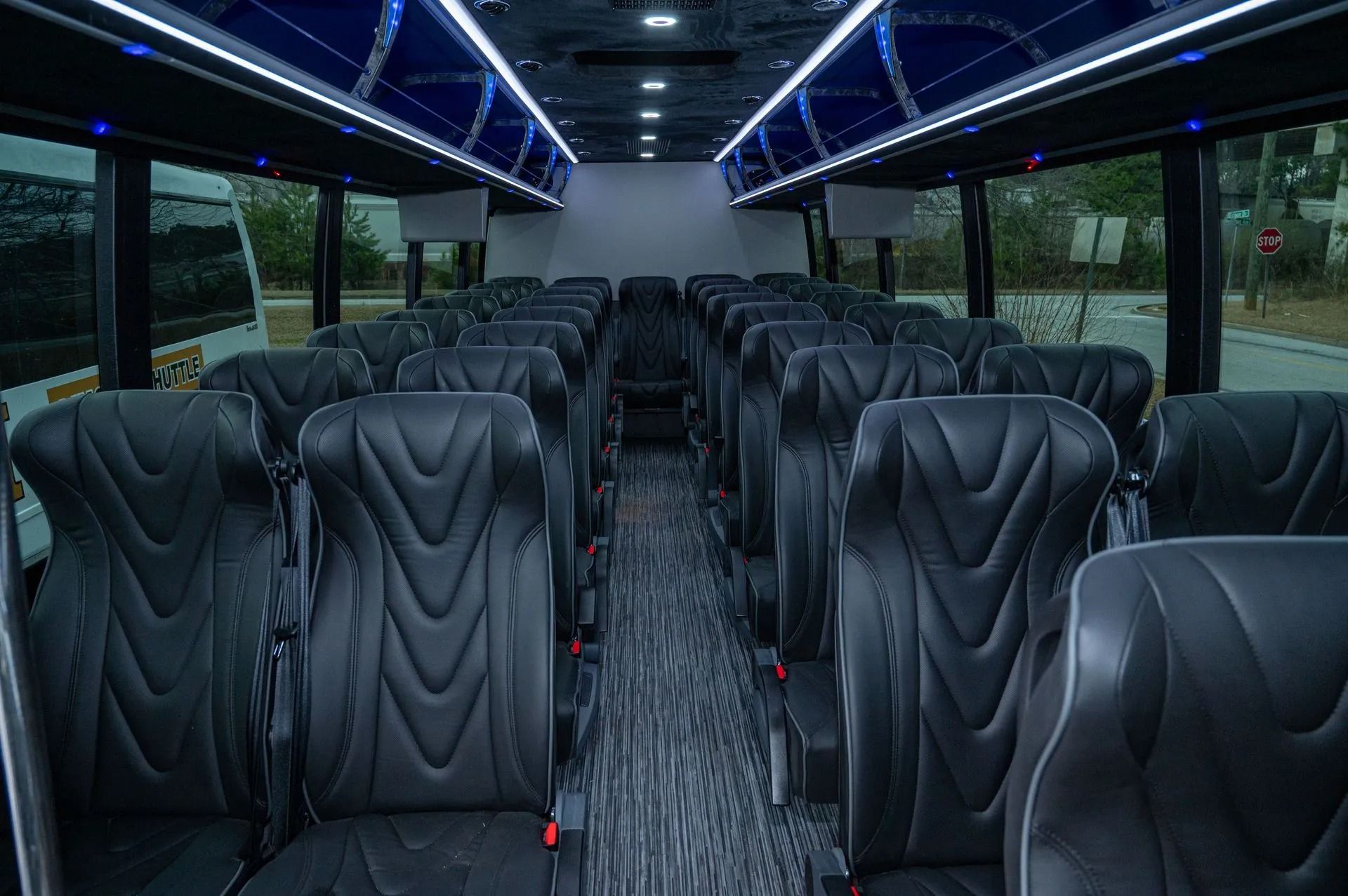The interior of a bus with rows of black leather seats, a grey patterned carpet, and blue ambient ceiling lighting.
