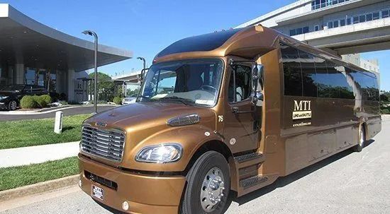A bronze MTI luxury shuttle bus parked outdoors on a sunny day in front of a modern building.