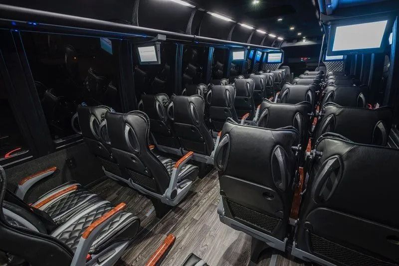 Interior of a luxury bus with rows of black leather passenger seats, wooden flooring, and mounted overhead screens.