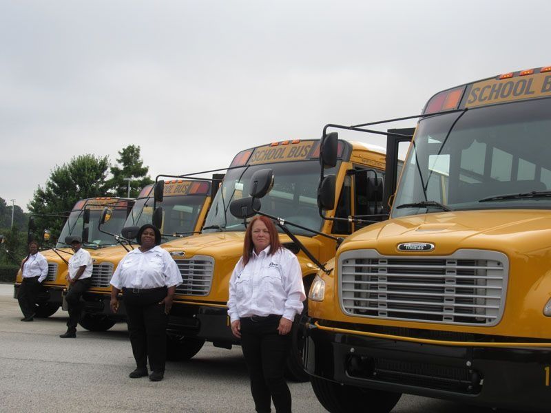 Four school bus drivers in white shirts and black pants stand in front of their parked yellow school buses.