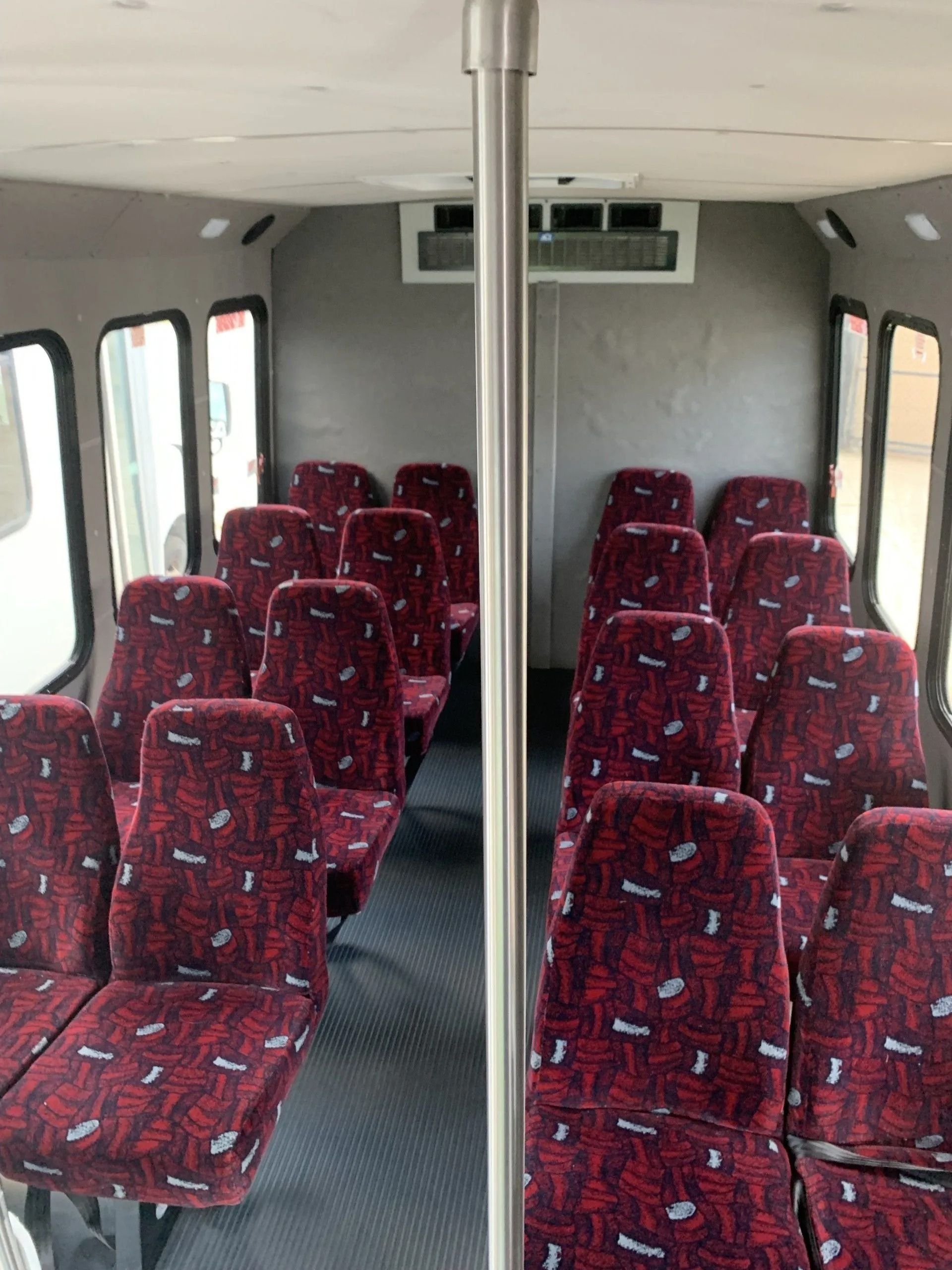 Inside a bus cabin with rows of red-patterned seats facing forward, divided by a central silver pole.