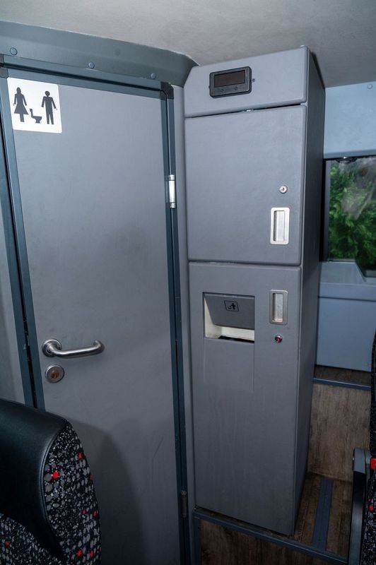 Gray restroom door beside a tall partition with signs and a toilet paper dispenser in a narrow hallway