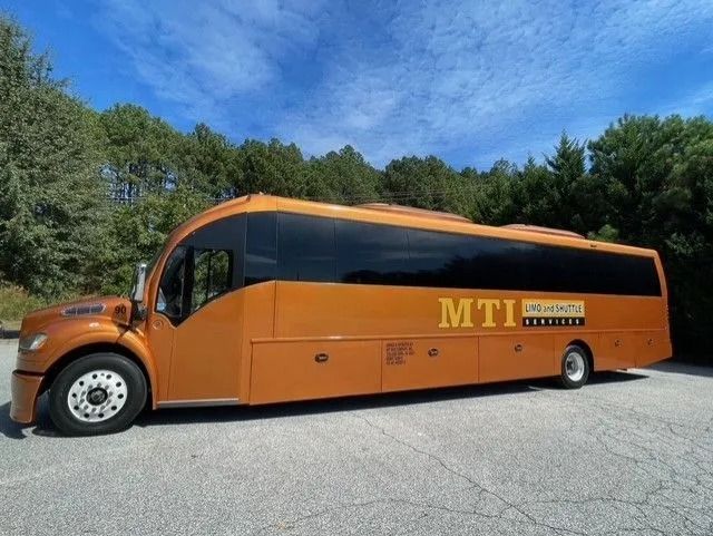 A copper-colored MTI motorcoach bus parked on an asphalt lot in front of a tree line under a bright blue sky.