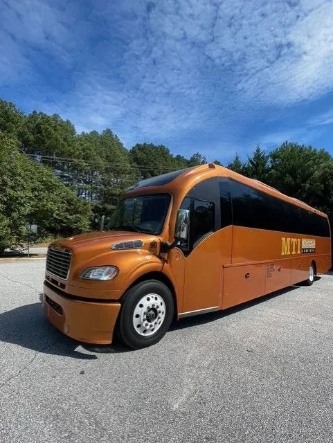 A metallic copper-colored shuttle bus parked outdoors under a blue sky, marked with the MTI logo on its side.