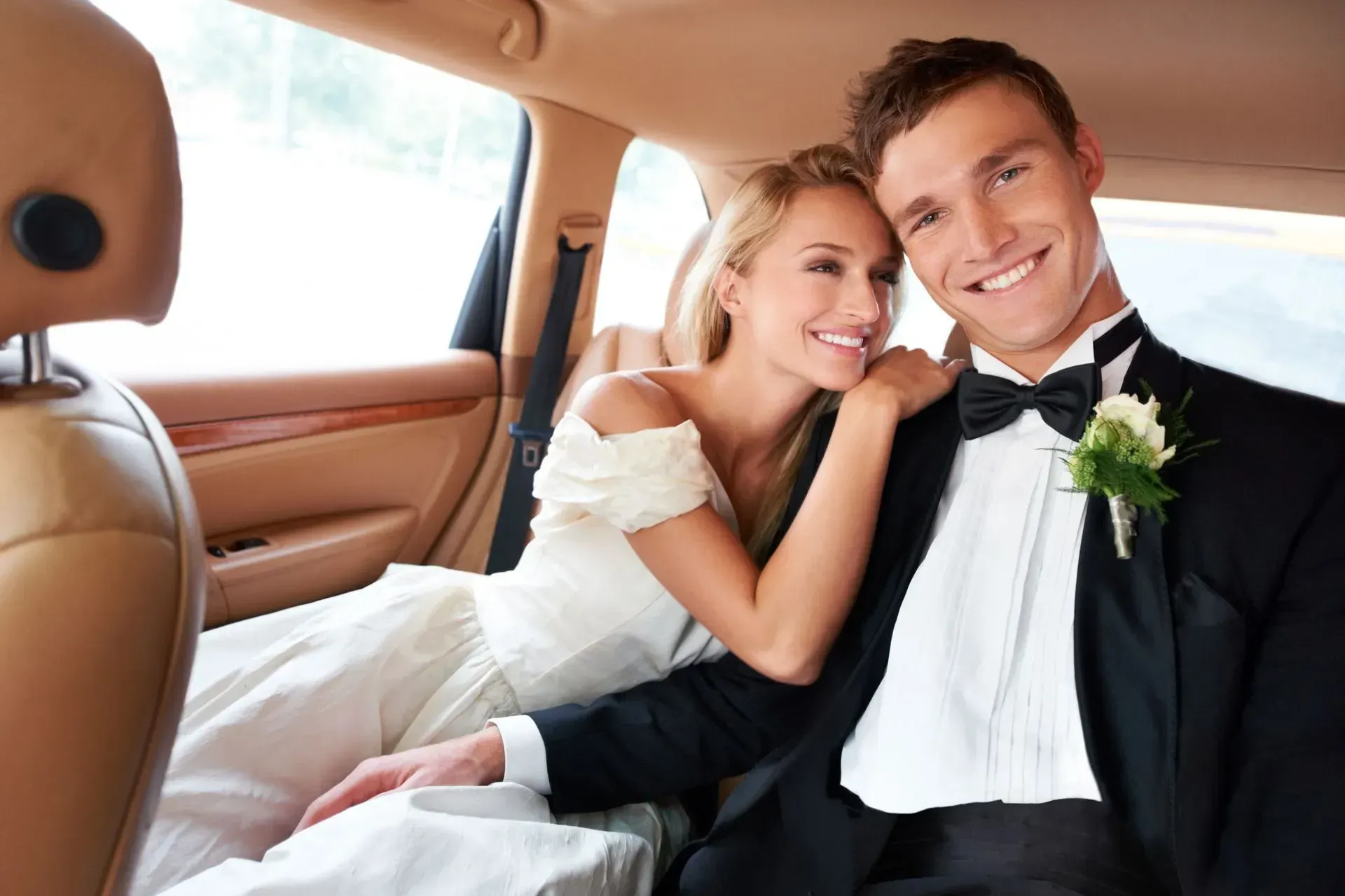 Newlywed couple smiling in a car, bride in white dress leaning on groom in black tuxedo