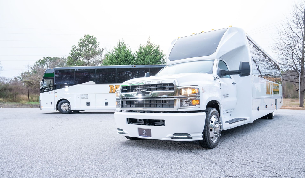 White charter bus and silver utility truck parked on a cracked lot with trees in the background