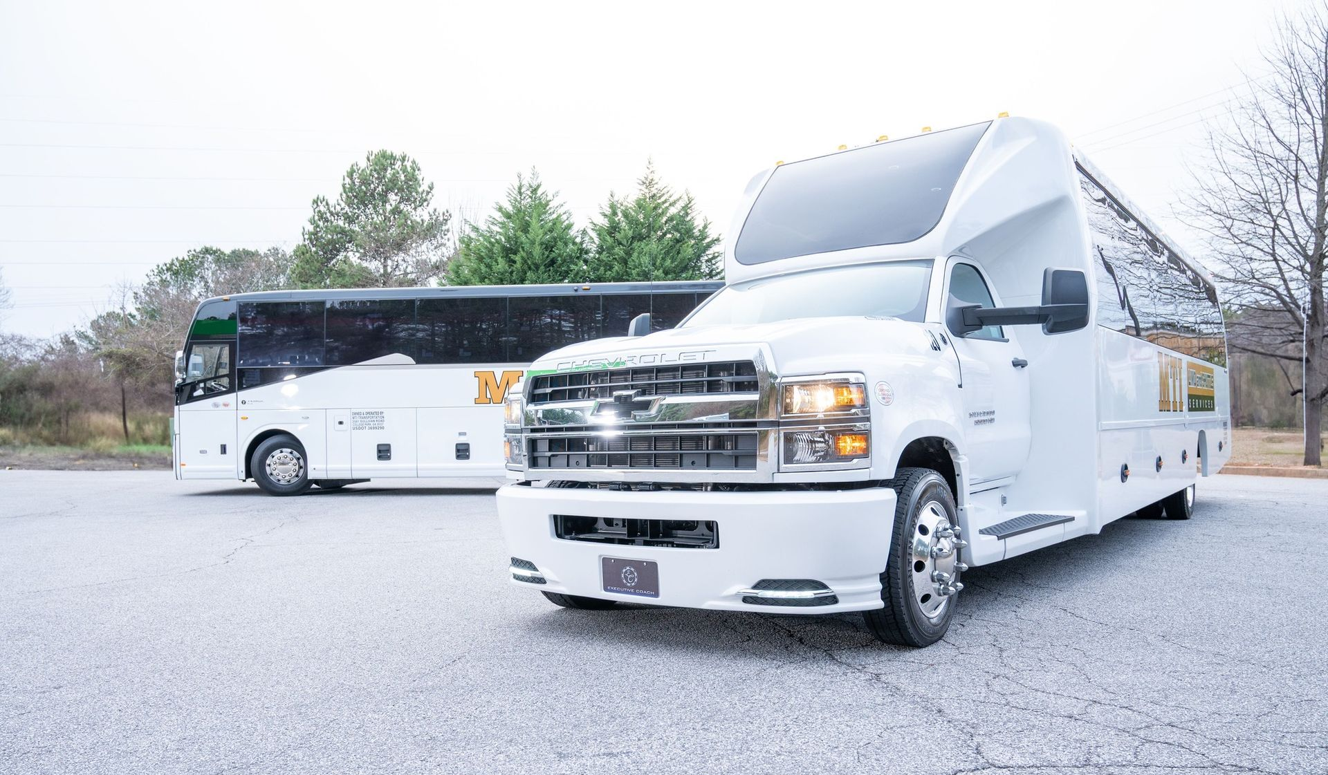 White charter bus and silver utility truck parked on a cracked lot with trees in the background