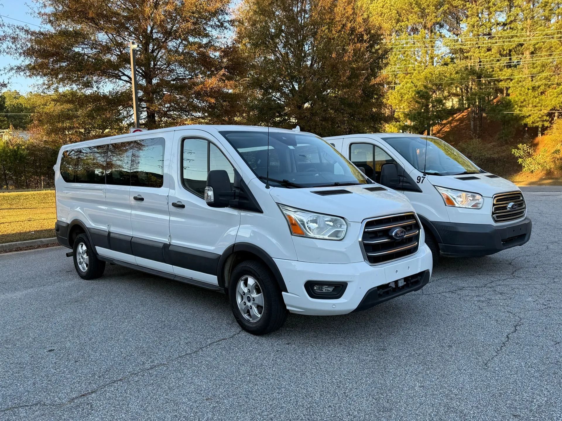 Two white Ford Transit passenger vans parked on an asphalt lot with trees in the background.