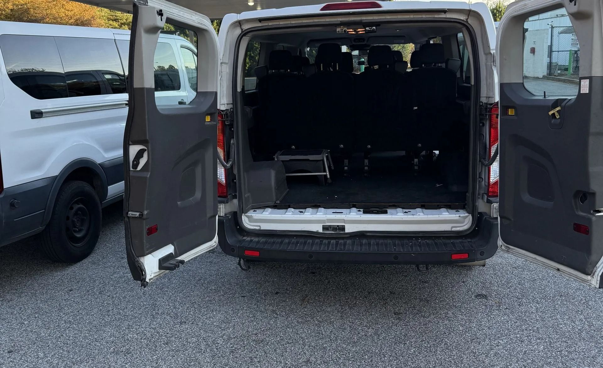 The rear view of a white van with its double back doors open, revealing an empty cargo or passenger area on a gravel lot.