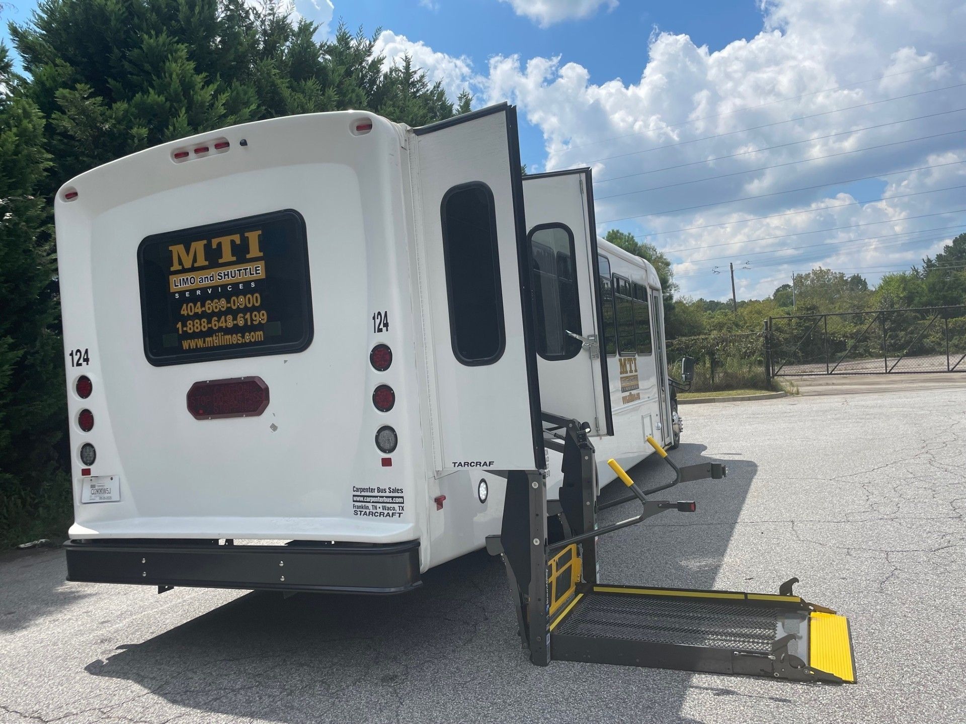 A white shuttle bus with a deployed wheelchair lift parked on a gravel lot under a cloudy blue sky.