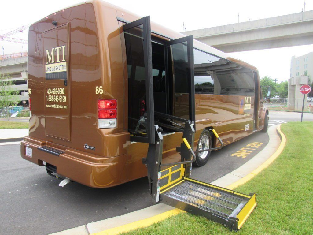 A brown shuttle bus parked at a curb with its side-mounted wheelchair lift fully extended to the ground.